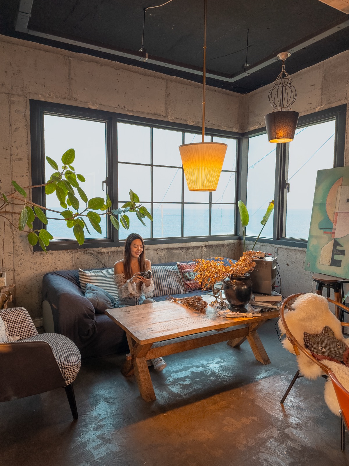 Woman sitting on a sofa in an industrial-style cafe with large windows overlooking the ocean at Cafe Rooftop in South Korea