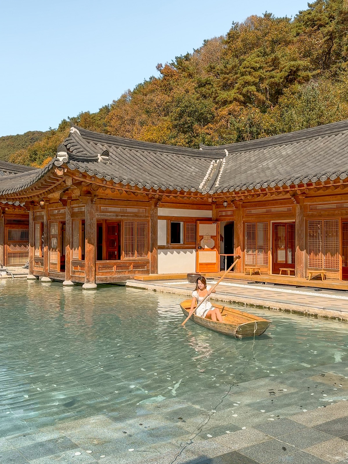 Woman rowing a wooden boat on a still pool in front of a traditional hanok building surrounded by autumn foliage at Cafe Tirol, South Korea