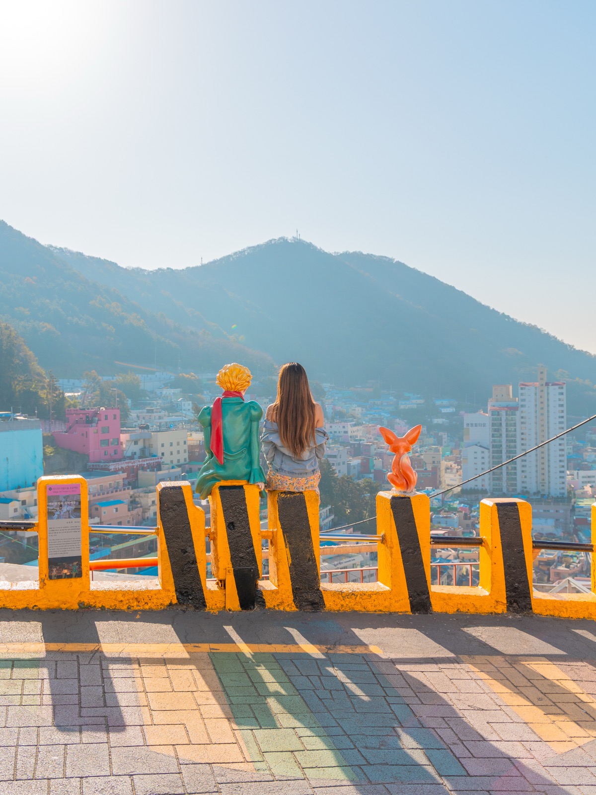 Woman sitting with Little Prince art installation overlooking the colorful hillside houses of Gamcheon Culture Village in Busan, South Korea