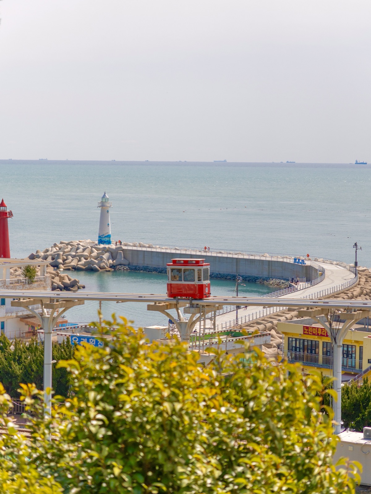 Red Sky Capsule cable car on elevated curving track along the Busan coast with a white lighthouse and ocean behind it