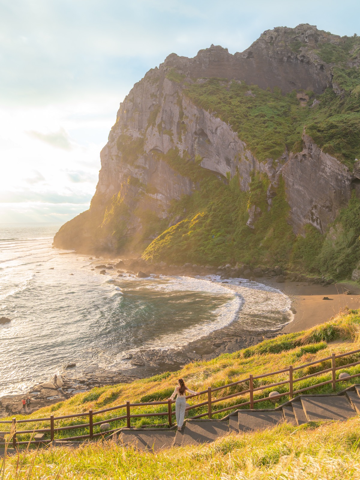 Woman standing on wooden steps overlooking dramatic coastal cliffs and a small beach cove on Jeju Island, South Korea