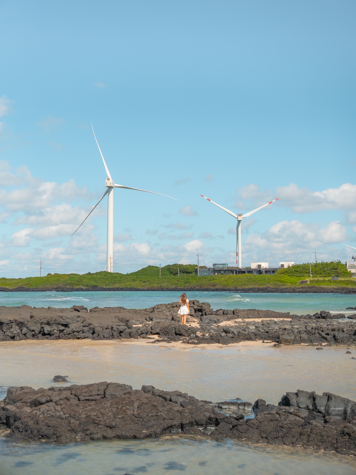 Woman standing on black volcanic lava rocks on a Jeju Island beach with two wind turbines and turquoise water in the background