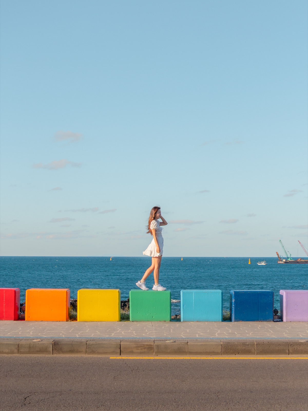 Woman standing on a row of rainbow-painted colorful blocks along the Jeju Island seafront with the blue sea behind her
