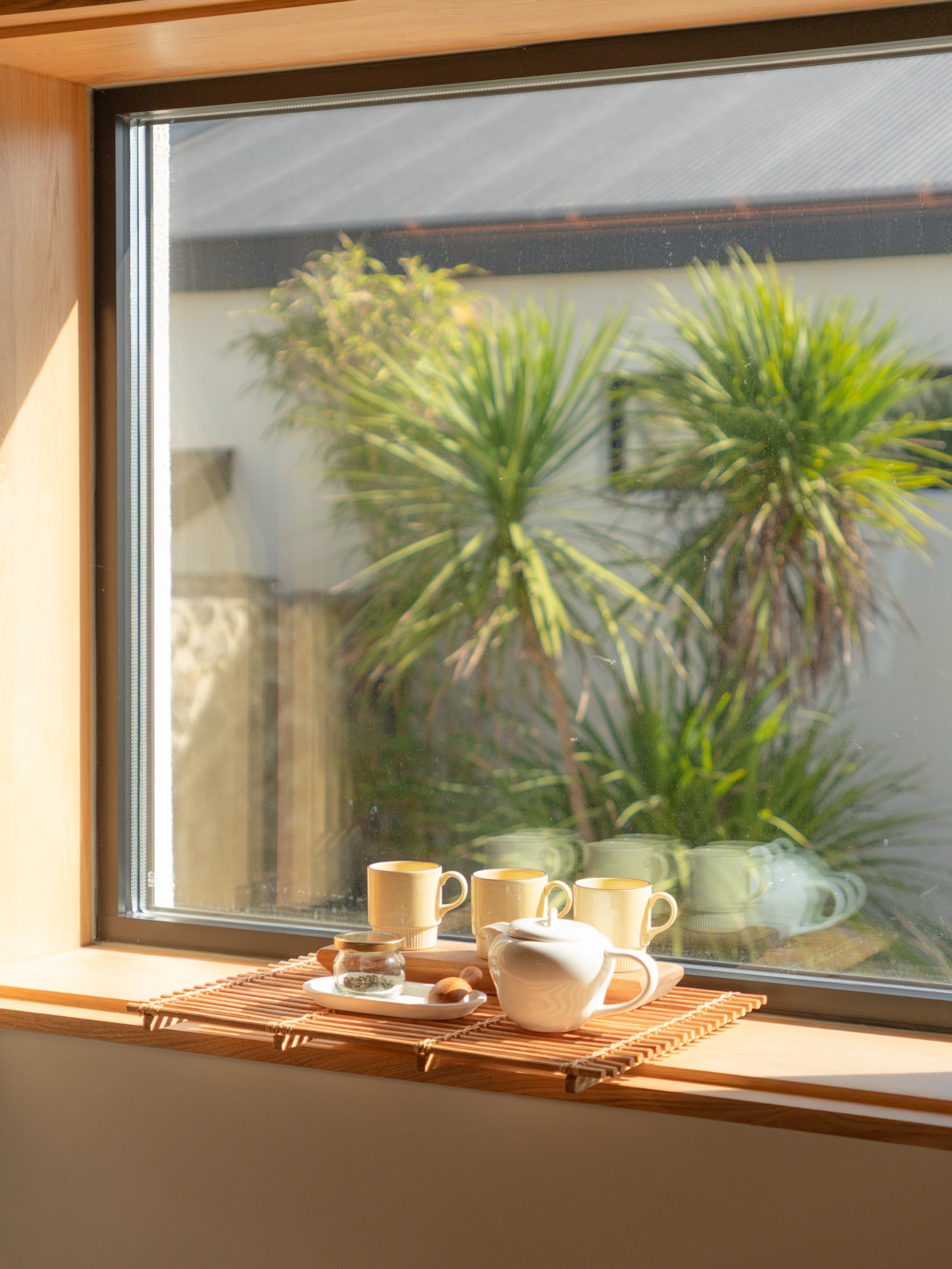 Yellow mugs and a white teapot on a bamboo mat by a sunlit window with palm trees outside at a hanok stay in Jeonju
