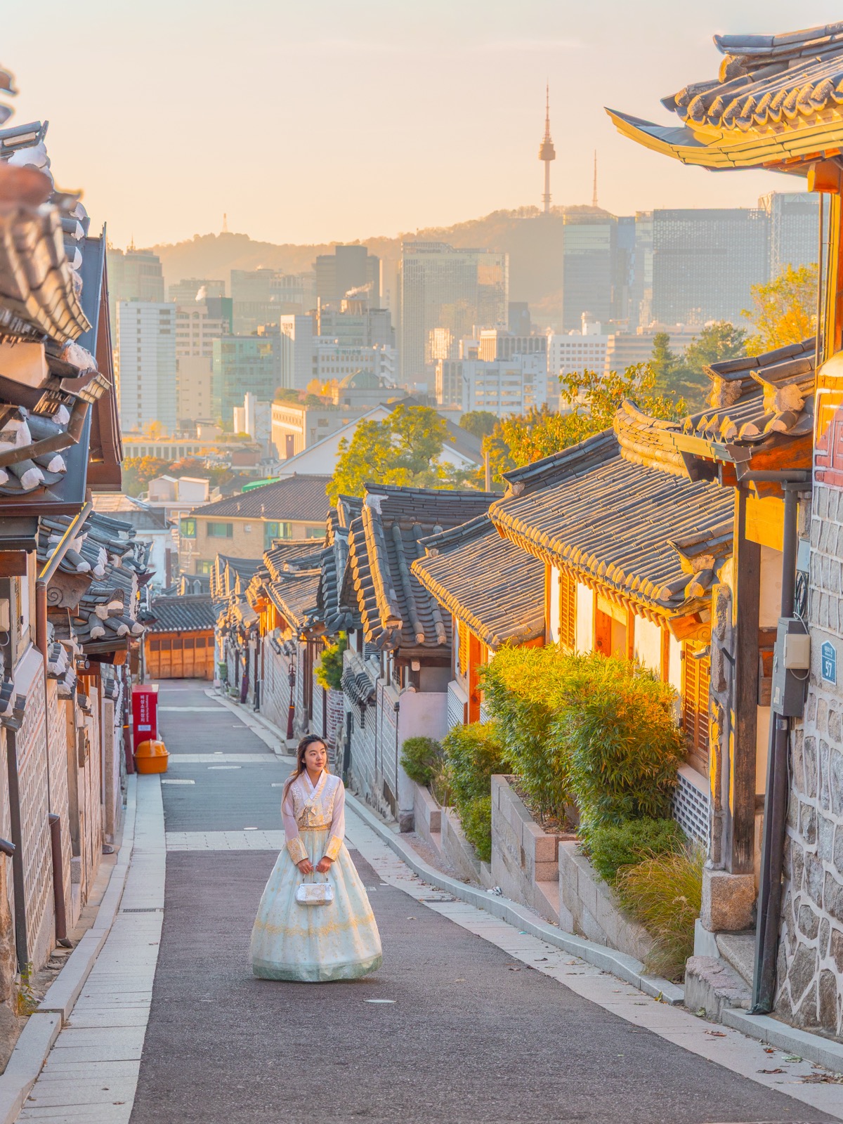 Woman in hanbok standing on the street of Bukchon Hanok Village at golden hour with N Seoul Tower visible in the background, Seoul South Korea